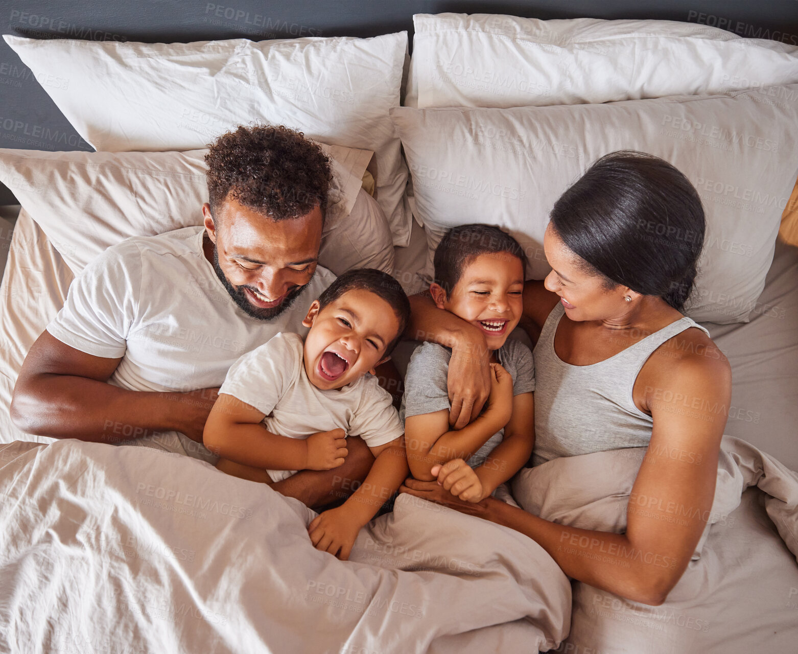 Buy stock photo Happy family bonding in bed together, playing and laughing while being loving and having fun. Young caring interracial parents sharing special moment of parenthood with two playful boys in bedroom
