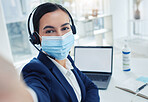 Customer support woman taking selfie on phone and working on laptop with headset and face mask. Receptionist or call center agent taking picture with smartphone camera at work during global pandemic.