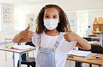 Thumbs up for education with covid, school student pleased with learning wearing a mask and showing support in a classroom at school. Portrait of a little girl protecting from virus during pandemic