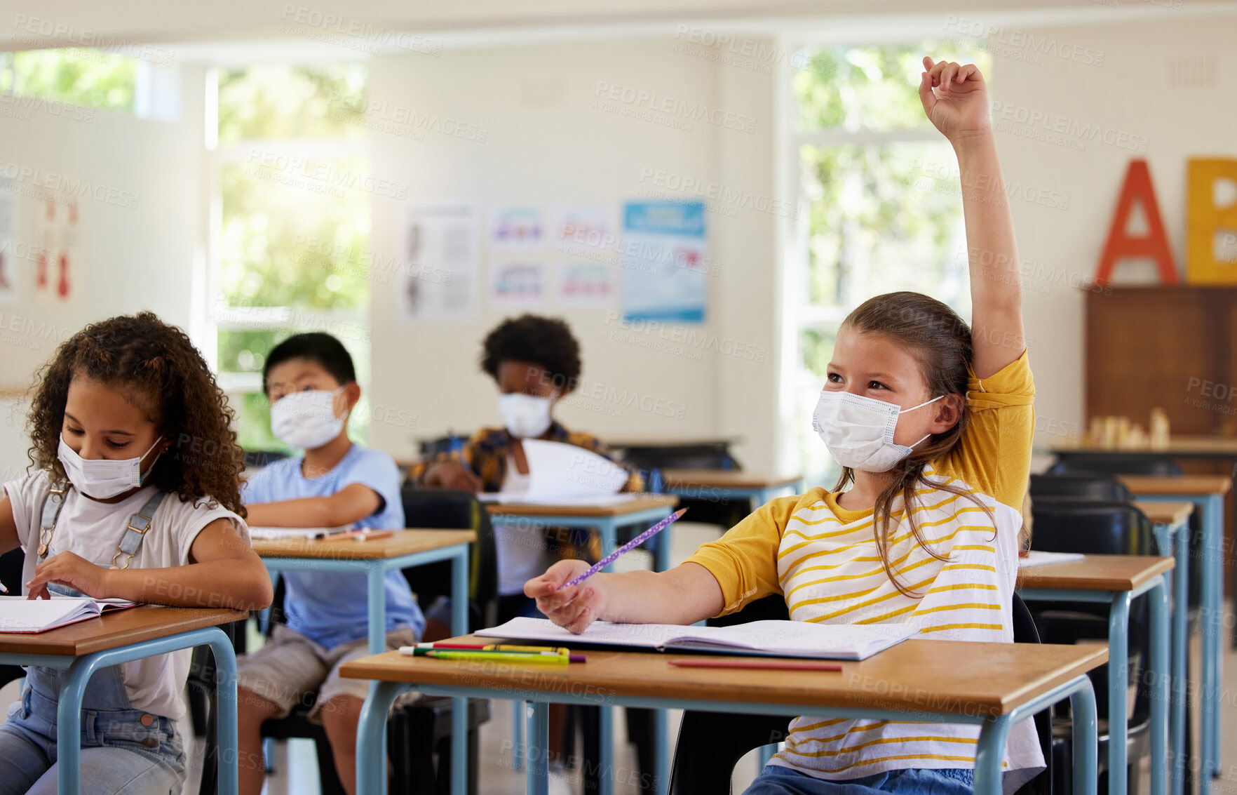 Buy stock photo Wearing face mask to protect from covid while learning in class, answering education question and studying with students in a classroom. Girl sitting at a desk and raising hand during a pandemic