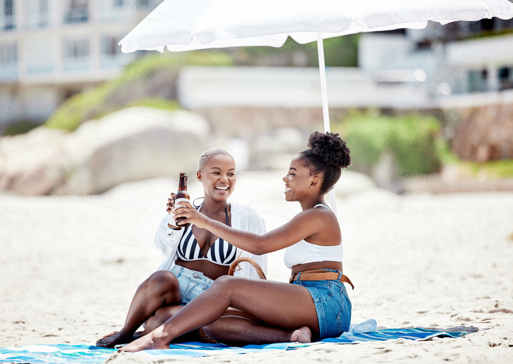 Buy stock photo Friends relaxing and drinking a beer on the beach while on vacation together during summer. Happy black women making a toast with alcohol while sitting by ocean while on holiday in south africa.