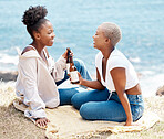 Friends drinking a beer on the beach while talking and relaxing on a summer tropical holiday. Happy black women having fun while on an ocean picnic with an alcohol drink enjoying their vacation.