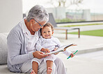 Senior woman, happy baby and children book reading of a grandmother spending quality time together. Elderly retirement of a old female about to read a fun kids story to a kid on a home patio 