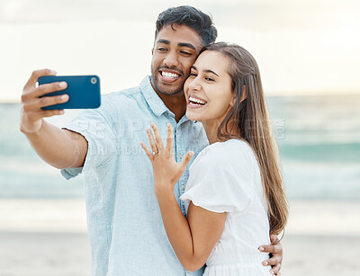 Buy stock photo Love, beach and couple after their engagement taking a selfie on a phone while on summer vacation. Happy woman showing off her ring after a proposal while taking picture with a man while on holiday.