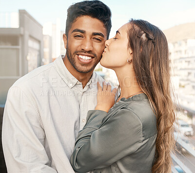 Buy stock photo Happy couple, love and kiss on cheek from girlfriend bonding with boyfriend standing on a city building balcony on a holiday. Portrait of indian man and beautiful woman in interracial relationship  