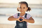 Girl at beach make heart sign with hands, happy and smile against blurred nature background. Young female child with expression of happiness, makes love icon with fingers by the ocean or sea