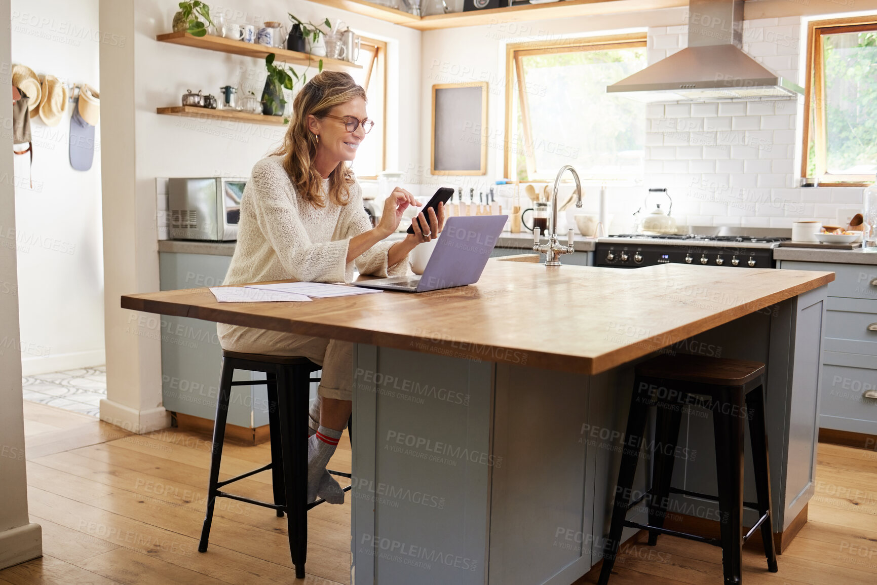 Buy stock photo Woman, phone and kitchen smile for social media, email or text while work online. Mature, lady and smartphone with laptop for communication in remote job on web, laughing at meme or joke on cellphone