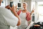 Senior couple cooking, smartphone taking portrait in kitchen and woman with v peace sign for social media, celebration of retirement or healthy lifestyle. Elderly people on cellphone with home photo