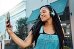 Phone, selfie and happy black woman in the street in the city with headphones listening to music, podcast or radio. Happiness, technology and african girl on a walk in the urban road on a smartphone.