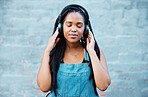 Black woman, headphones and music with a girl feeling calm, relax and content listening to a podcast standing outside against city or urban wall. African female relaxed during mindful audio streaming