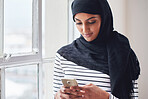 Muslim woman using smartphone texting on mobile phone browsing messages wearing hijab headscarf standing by window