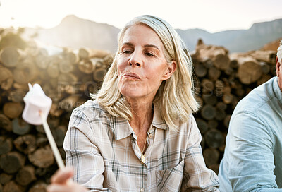 Buy stock photo Senior woman eating marshmallow with regret, relaxing by the campfire after her mountain adventure in the outdoors nature. Female enjoying wellness lifestyle, resting after hiking trip with friends