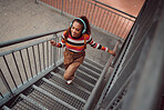 Girl with headphones walking up stairs in the city, phone in hand and listening to music. Happy black woman student streaming song, track or radio on smartphone at university or college in urban town
