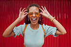 Funny, happy portrait of black woman face and smile with her tongue out with designer fashion sunglasses on red background. Girl with crazy comic expression, modern hair style and trendy accessories 