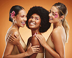 Beauty, flowers and women in makeup, diversity and models on orange studio background. Smile, skincare and happy, young and elegant female friends posing together with orchids after spa treatment.
