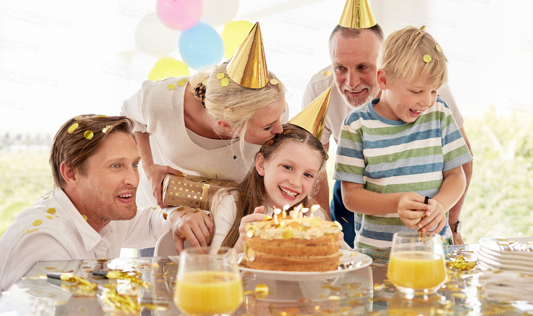 Buy stock photo Girl, birthday cake and family celebrating a children event with food and a party outdoor. Happy smile of kids, father and mom kiss to celebrate youth and kid event with happiness, love and confetti