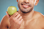 Man, hands and holding apple on blue background in studio for health, wellness or immune support. Zoom, smile or happy personal trainer with diet fruit for weight loss, wellness or energy for workout