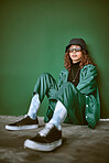 Fashion, style and clothes with a model black woman sitting on the floor against a green background. Portrait, glasses and fashionable with a young female posing to promote trendy or edgy clothing