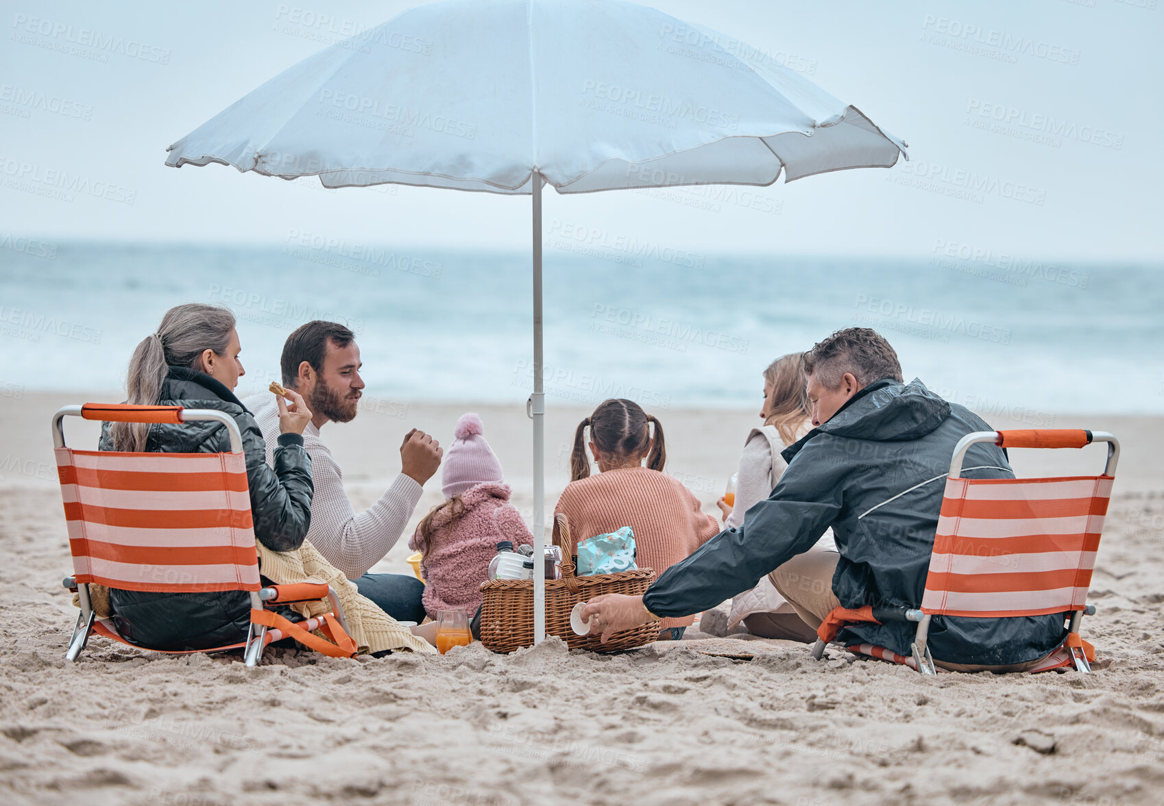 Buy stock photo Family, beach food and relax on holiday, vacation or trip outdoors on sandy seashore. Back view, bonding and grandparents and mom, dad and kids enjoying quality time together under beach umbrella.