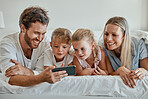 Happy family with kids, phone and lying on bed in home, mother, father and girl children watching video together. Love, fun and family with dad, mom and daughters on smartphone video call in bedroom.