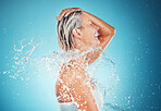 Splash, water and mature woman shower, skincare and cleaning for health, hygiene and wellness against a blue studio background. Happy elderly female beauty, shower and wash body in bathroom grooming
