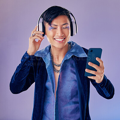 Buy stock photo Headphones, studio and gay man with makeup listening to music on the internet with smartphone. Cosmetics, lgbtq and happy guy streaming audio, podcast or radio on phone isolated by purple background.