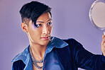 Asian man with glitter makeup, mirror for creative beauty and lgbt gender performance in fantasy art. Purple background in studio, punk fashion jewellery in Seoul and proud of unique fluid identity