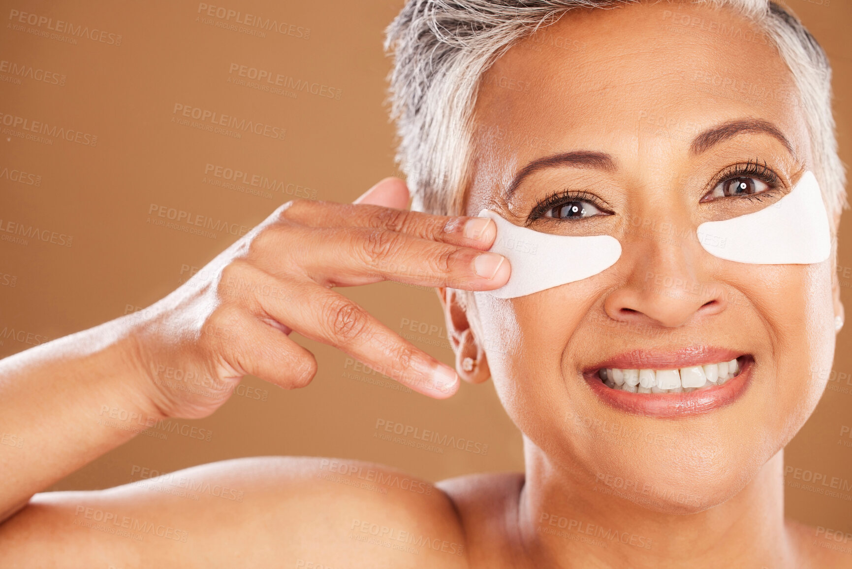 Buy stock photo Beauty, skincare and senior woman with eyepads on face for a natural facial routine in studio. Cosmetic, wellness and portrait of elderly lady with skin treatment product isolated by brown background