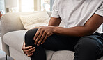 Knee pain, hand of a man and injury while sitting on home living room sofa for rest and relaxation. Ache, painful leg joint and african american man touching his knees for a strain muscle 