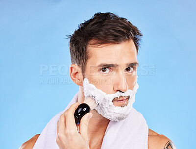 Buy stock photo Face, shave and grooming with a man model shaving his skin for hair removal in studio on blue background. Portrait, brush and hygiene with a handsome young male in the bathroom for a morning routine