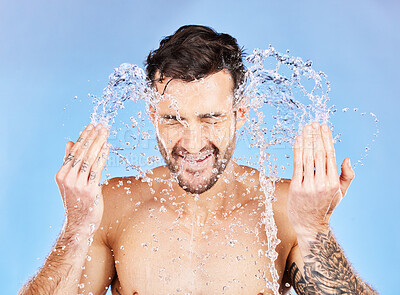 Buy stock photo Face, water and cleaning with a man model splash in studio on a blue background for hygiene. Skincare, water splash and hydrate with a handsome young male washing his skin in the bathroom for care