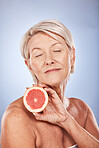 Face, skincare and senior woman with grapefruit isolated on a blue studio background. Happy, elderly and female model with eyes closed holding fruit for natural facial care, healthy skin or vitamin c