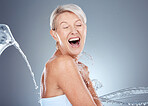 Water splash, health and portrait of senior woman shocked and surprised on gray background. Skincare, wellness and beauty, a happy mature lady splashed with water with happy smile on face in studio.