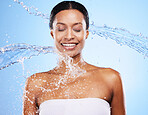 Woman, water splash and shower with body cleaning and smile with healthy skincare in blue studio background. Clean, face and water splash with an attractive young female cleaning in the bathroom