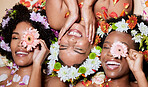 Top view, beauty and black women with flowers for skincare in studio on brown background. Makeup aesthetics, organic cosmetics and group, friends or female models with floral plants for healthy skin
