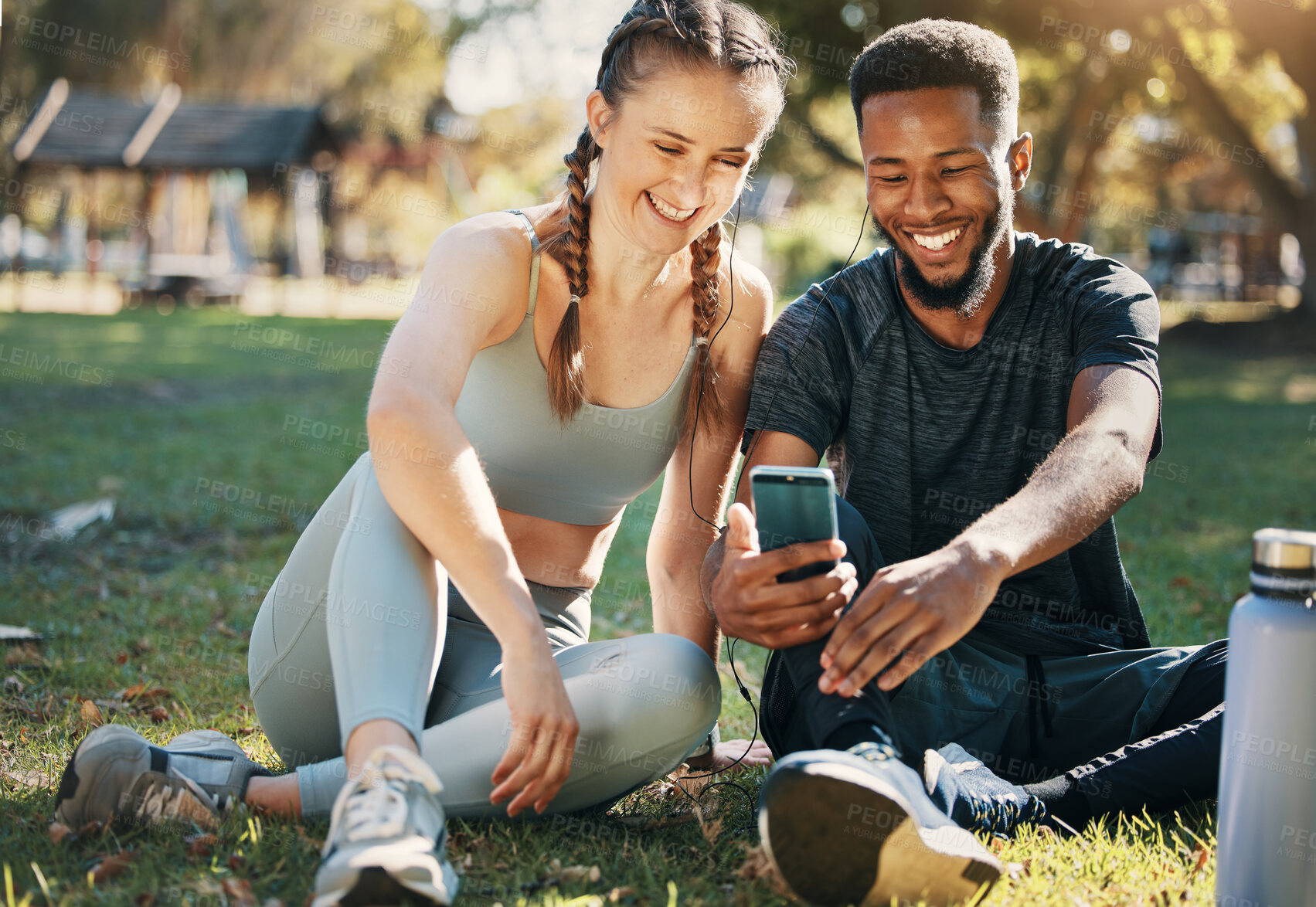 Buy stock photo Phone, earphones and couple on an outdoor exercise while watching funny video on social media. Fitness, sport and interracial man and woman listening to music together relaxing on grass after workout