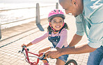 Black girl, learning and bike ride with dad at ocean promenade with smile, helmet or happy in sunshine. Kid, father and bicycle in training, safety or help for childhood development together outdoor