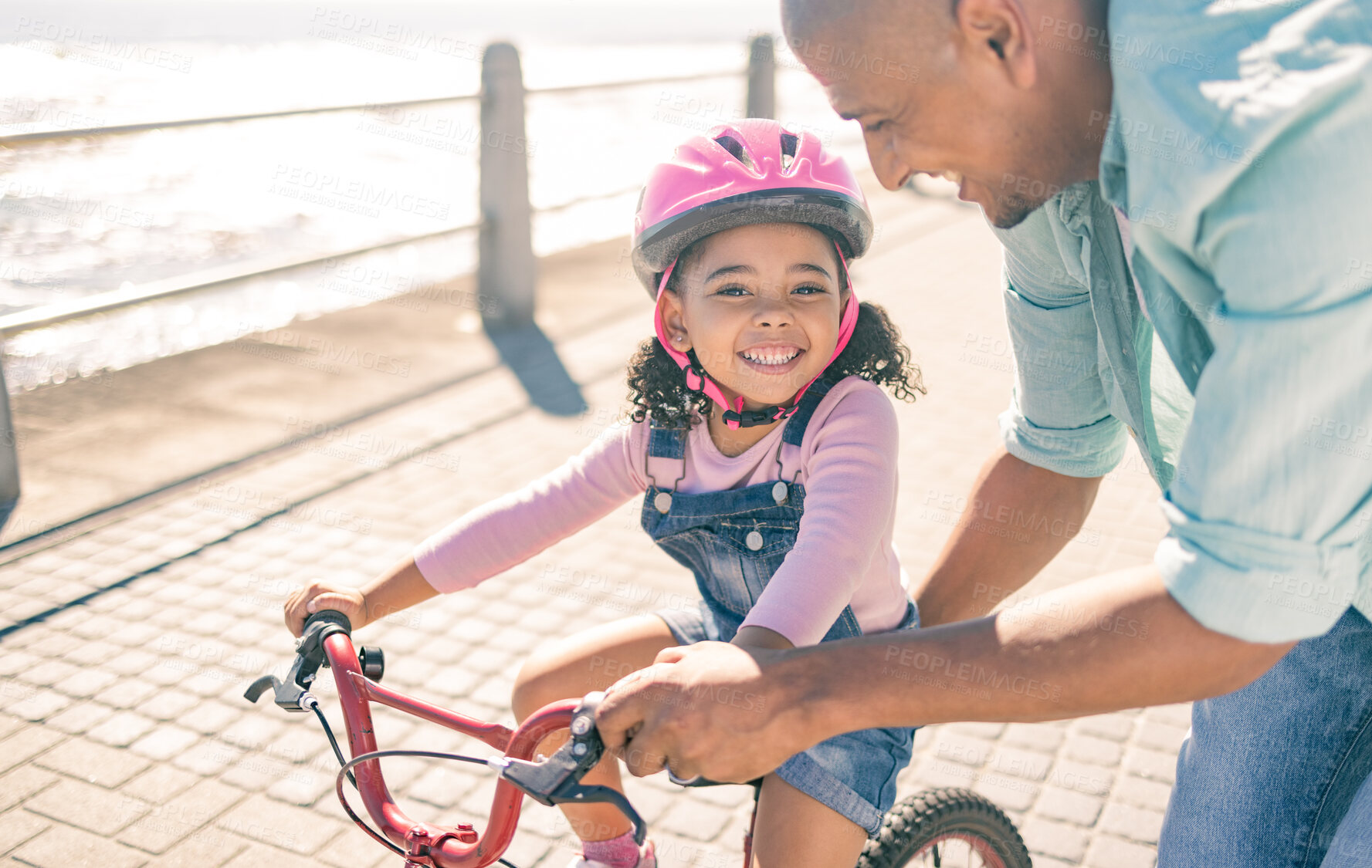 Buy stock photo Black girl, learning and bike ride with dad at ocean promenade with smile, helmet or happy in sunshine. Kid, father and bicycle in training, safety or help for childhood development together outdoor