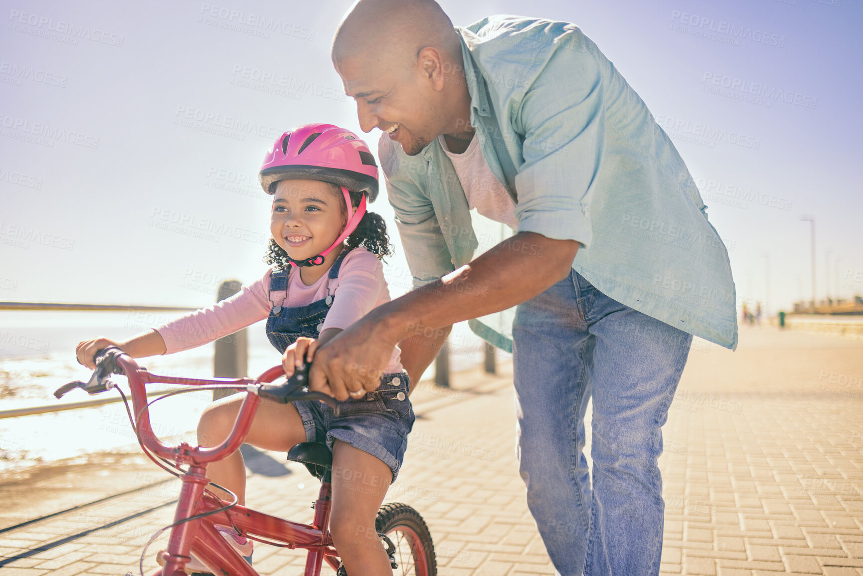 Buy stock photo Black girl, bike and learning with dad, smile and park in sunshine, safety or happiness by sea. Father, kid and bicycle for summer training, childhood development and outdoor in street with helmet