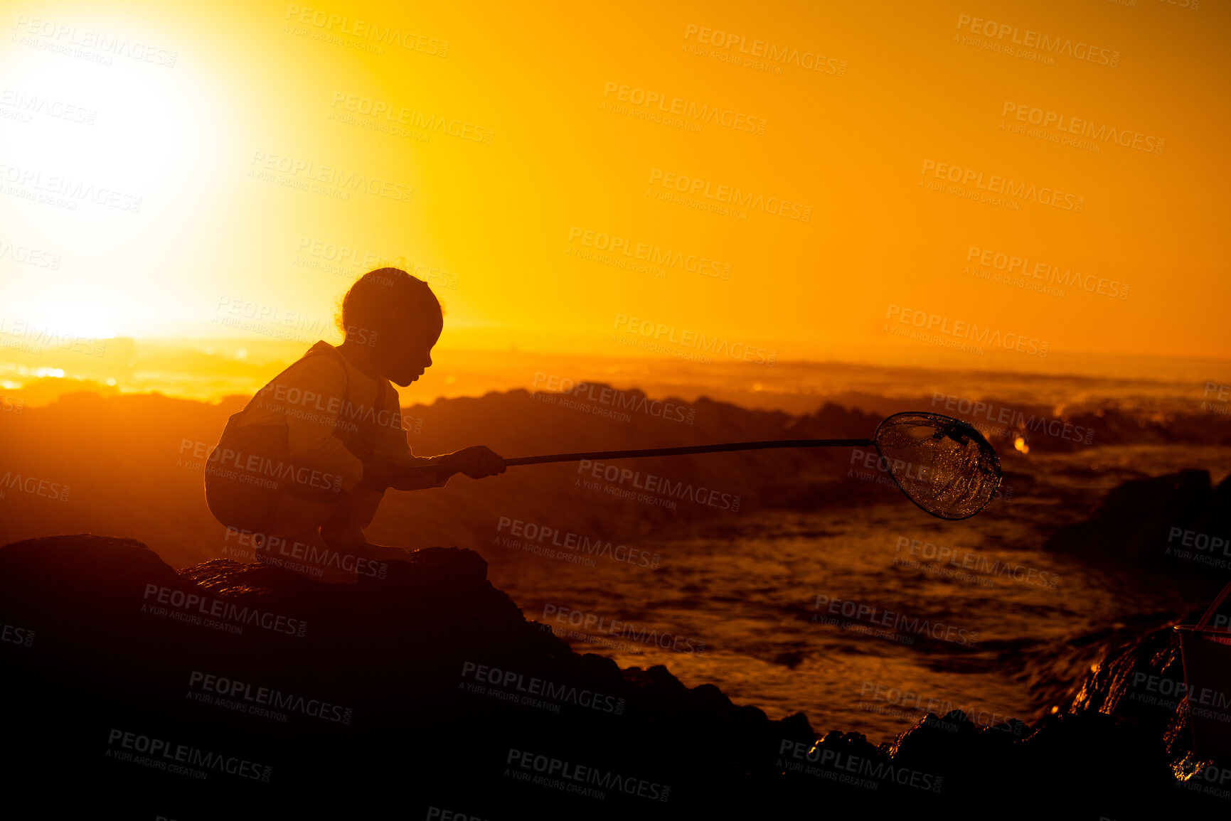 Buy stock photo Sunset, silhouette and child on a rock at the beach playing or fishing with net in the water. Travel, seaside and girl kid in nature by the ocean in the evening to play or have fun while on vacation.