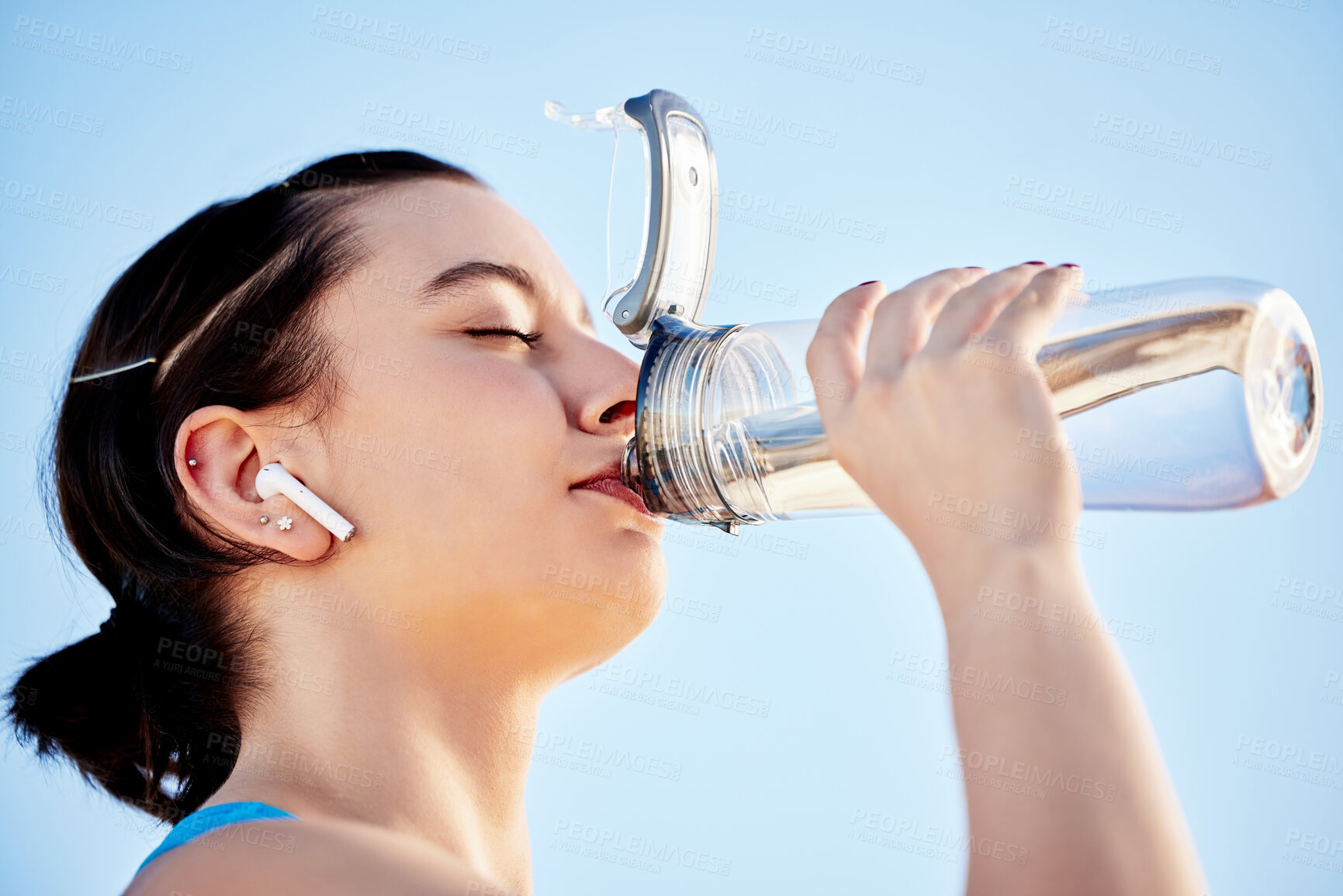 Buy stock photo Drinking water, fitness and music with a sports woman hydrating during a run on a blue sky background outdoor. Wellness, health and exercise with a female runner or athlete taking a drink to hydrate