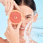 Face, water splash and woman with grapefruit, shower or skincare with happy smile, vitamin c or cleaning body. Female model, fruit or wellness in blue studio background with skin care or healthy skin