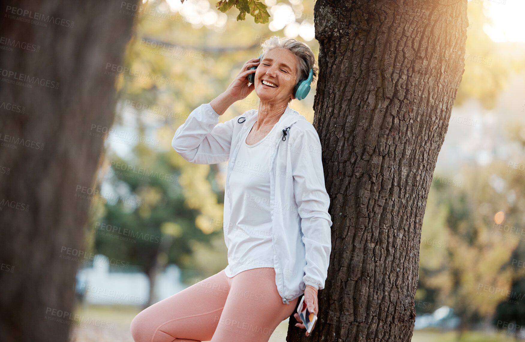 Buy stock photo Mature woman, headphone and music in a forest for exercise break in a park to rest by a tree. Workout, training and senior active female listening to cellphone audio song in a headset in a garden