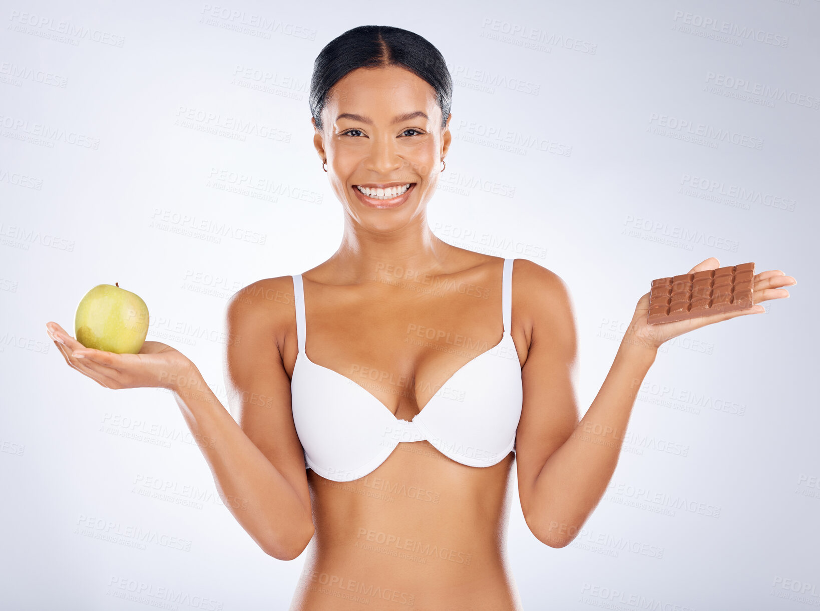 Buy stock photo Black woman, health and food choice for diet, nutrition or skincare vitamins against a grey studio background. Happy woman smiling in bra holding apple and chocolate in weight loss or body care plan