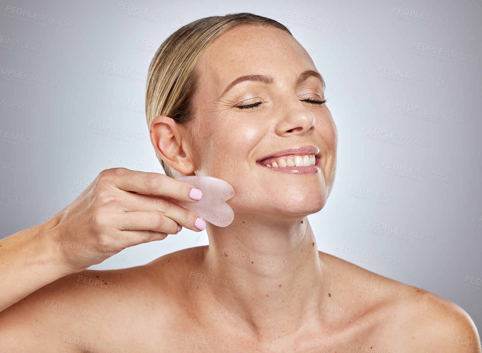 Buy stock photo Gua sha, skincare and woman with face massage, self care product and spa wellness against a grey studio background. Beauty facial, smile and model massaging skin for anti aging and natural glow