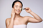 Face, beauty and skincare with a model asian woman using a facial roller on her skin in studio on a gray background. Massage, portrait and wellness with an attractive young female holding a product