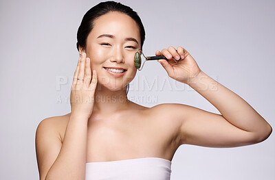 Buy stock photo Face, beauty and skincare with a model asian woman using a facial roller on her skin in studio on a gray background. Massage, portrait and wellness with an attractive young female holding a product