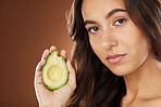 Face portrait, skincare and woman with avocado in studio on a brown background. Beauty, organic cosmetics and young female model with fruit, product or food for healthy diet, nutrition and vitamin c.