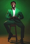 Fashion, trendy and black man model in a suit on chair in the studio with elegant, fancy and stylish outfit. Happy, smile and portrait of African male with formal clothes isolated by green background