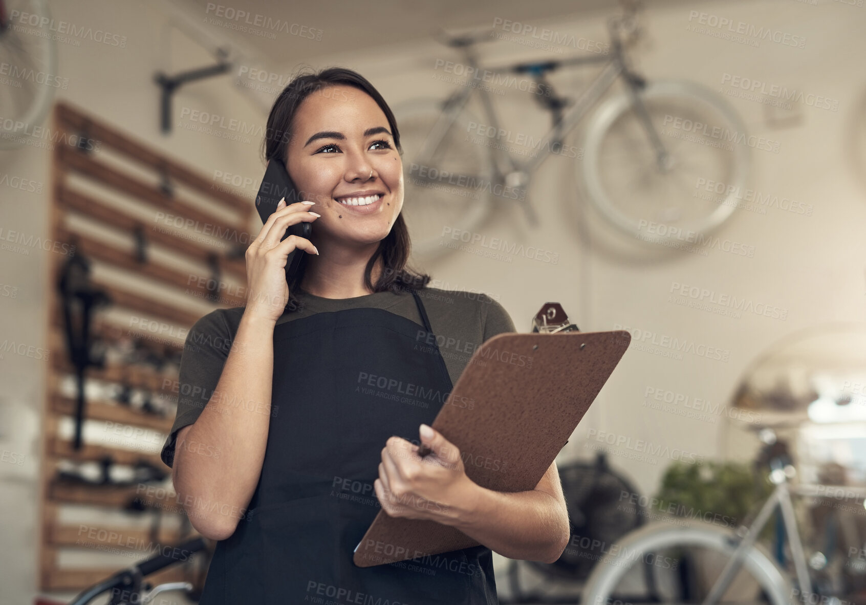 Buy stock photo Shot of an attractive young woman standing alone in her bicycle shop and using her cellphone while holding a clipboard